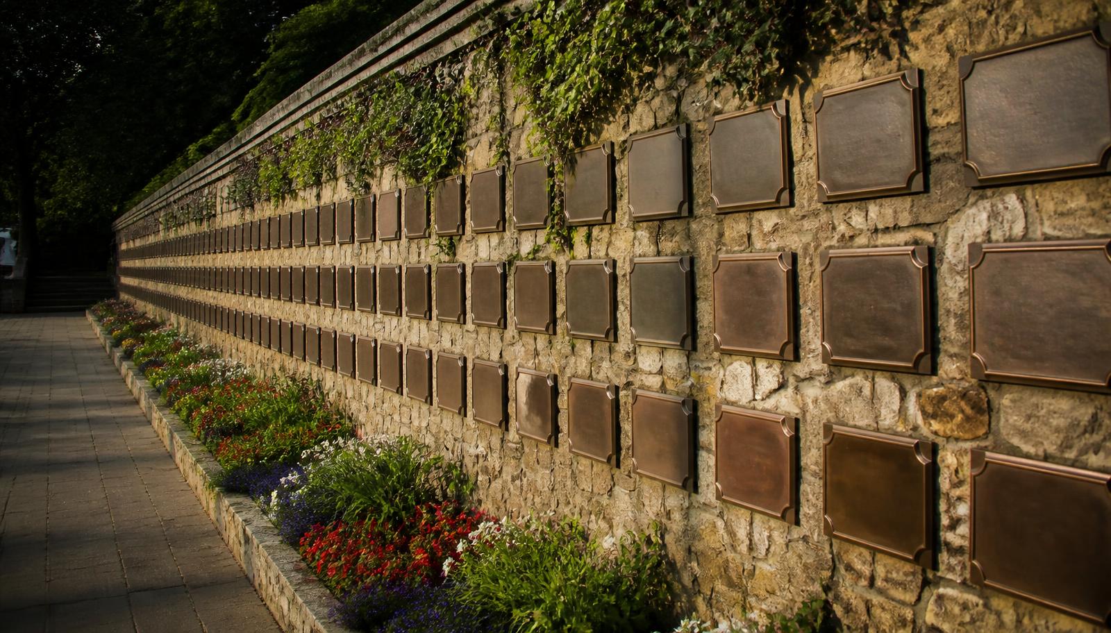 Long stone memorial wall lined with bronze remembrance plaques and flower beds